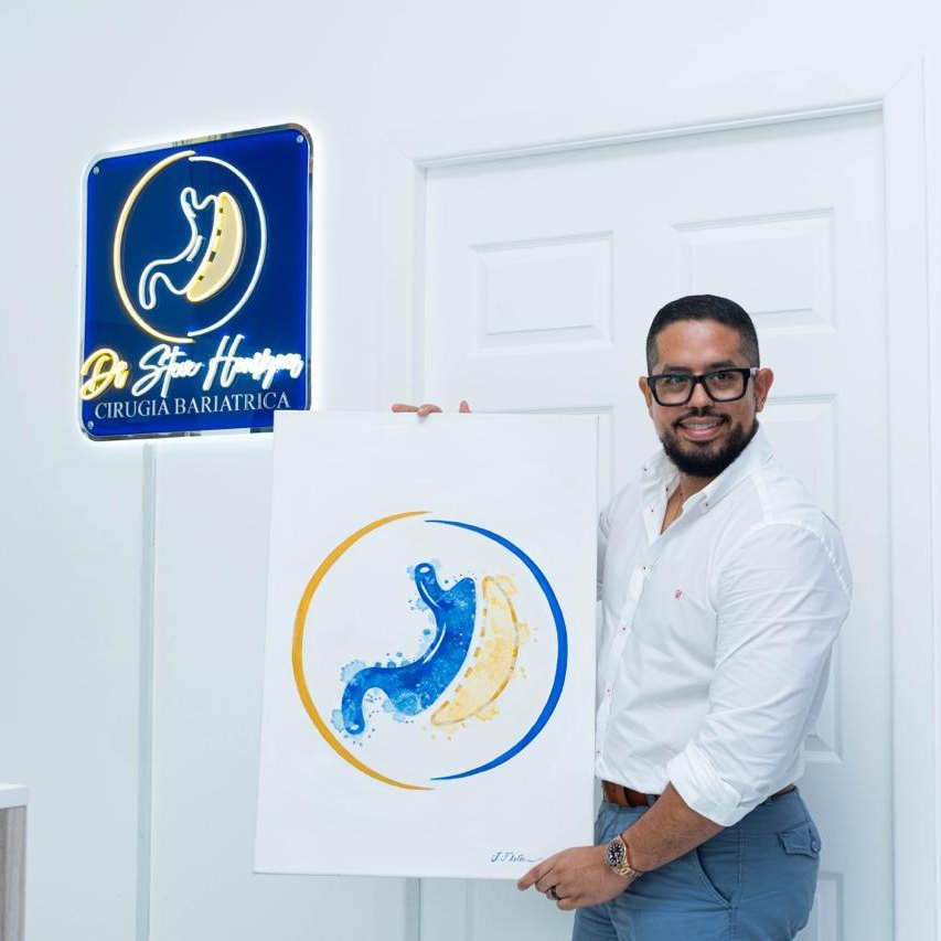 El Dr. Steve Henríquez en su clínica, junto al letrero con su logotipo y un cuadro con el mismo símbolo
